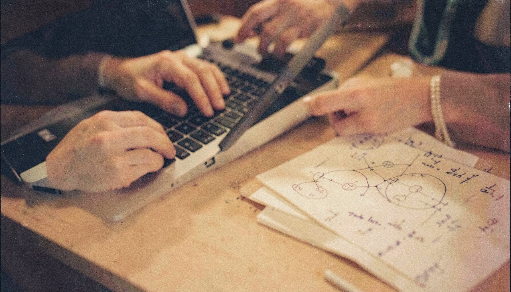 An analog film photograph with subtle grain depicting a close-up of hands typing on a laptop keyboard beside a notebook with handwritten notes, warm desk-lamp light across the scene.