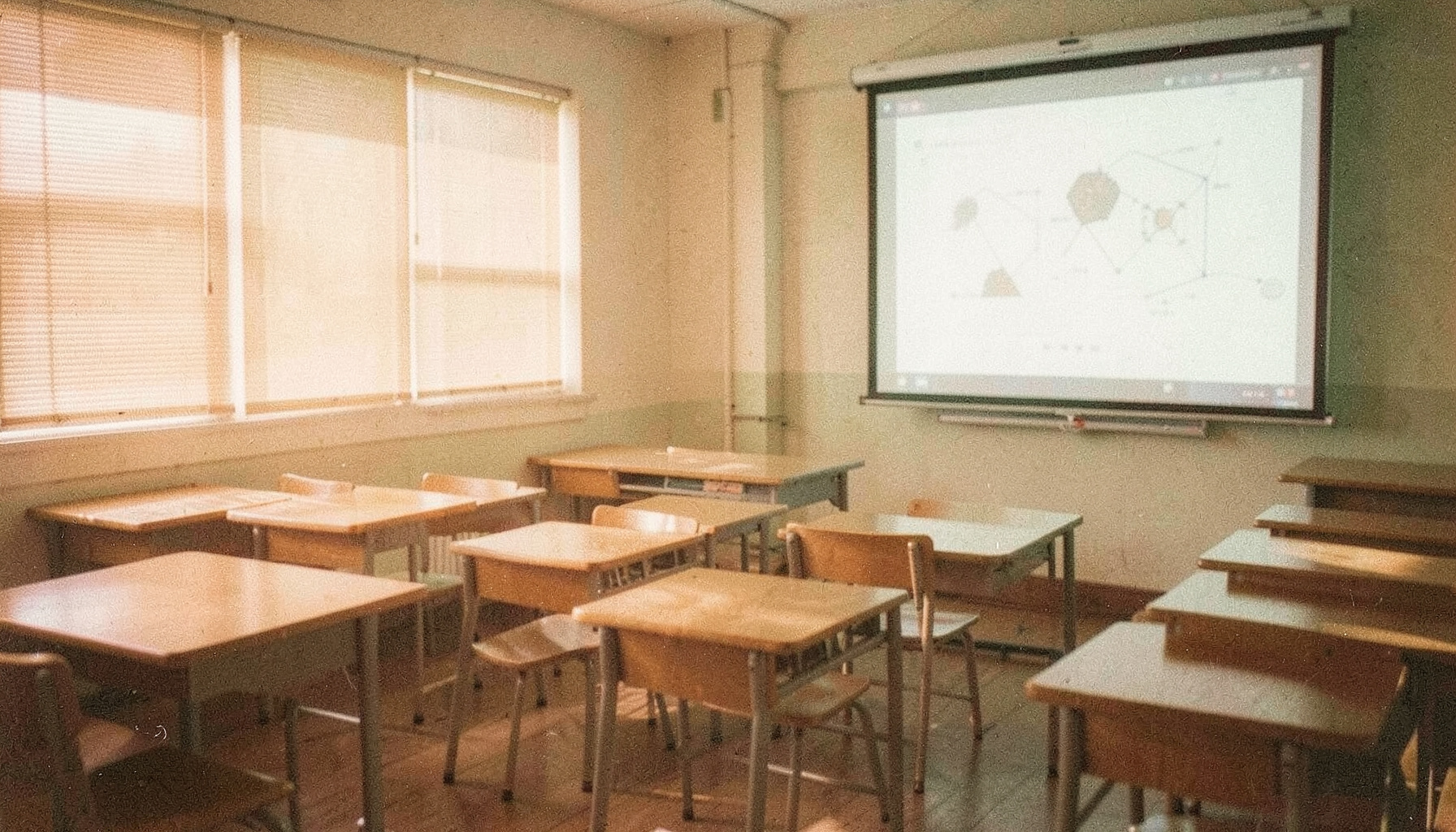 A warm analog film photograph with visible grain showing a quiet classroom with wooden desks, a projector screen displaying a presentation, and soft afternoon light falling through windows.
